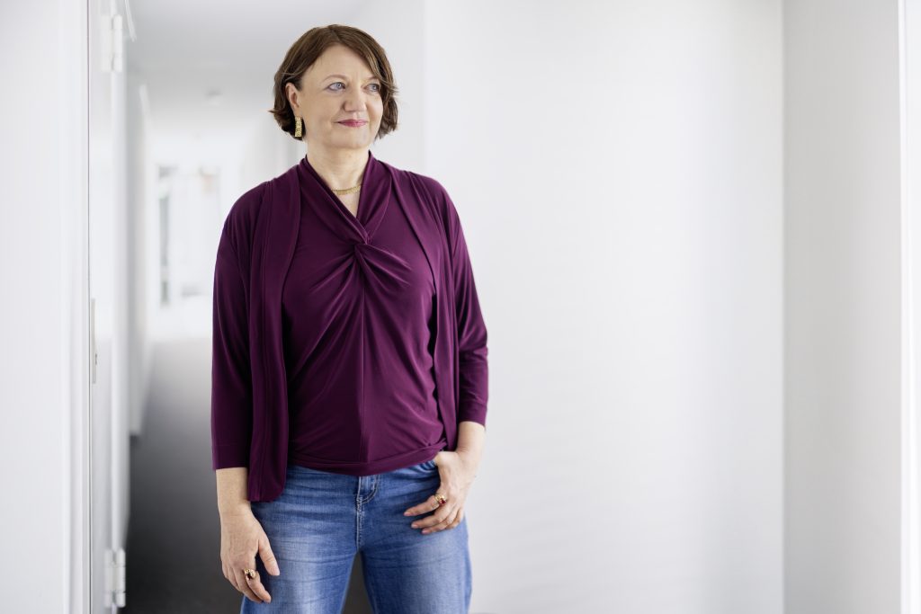 Foto der Autorin Eleonora Hummel stehend vor einer weißen Wand. Sie trägt ein weinrotes Oberteil und eine Jeans.