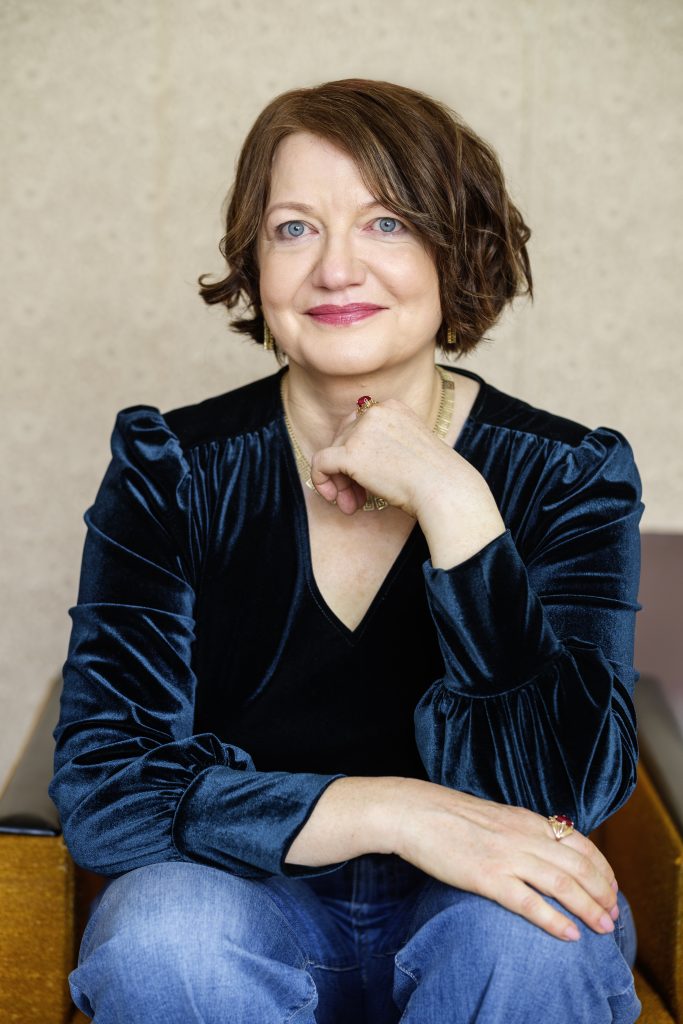 Porträtfoto der Autorin Eleonora Hummel sitzend vor einer Wand. Sie trägt eine Jeans und ein grünes Oberteil.